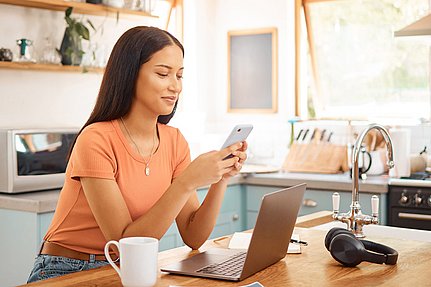 Eine junge Frau sitzt am Küchentisch und lächelt in ihr Smartphone. Vor ihr stehen ein Laptop, eine weiße Tasse und Kopfhörer. Die Küche ist hell und modern, mit Holzregalen und Sonnenlicht, das durch das Fenster hereinströmt.