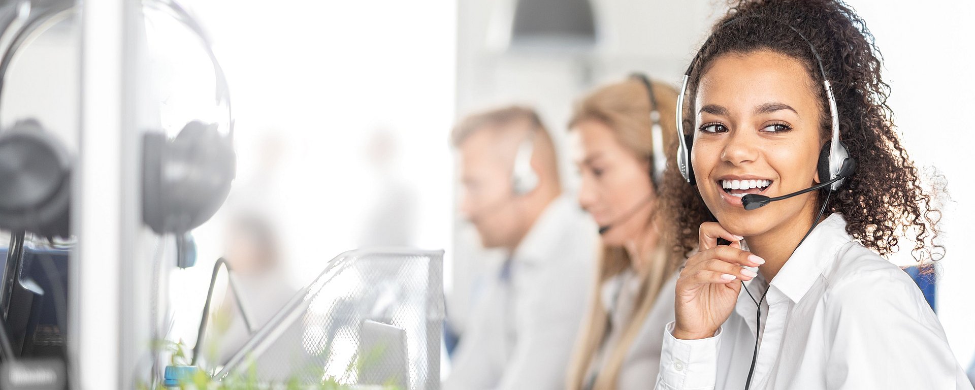 Eine lächelnde Frau mit einem Headset arbeitet in einem Callcenter, im Hintergrund sind ihre Kollegen unscharf zu sehen.