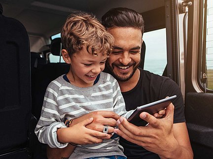 Ein lächelnder Mann und ein Kind sitzen in einem Auto und schauen sich gemeinsam ein Tablet an.