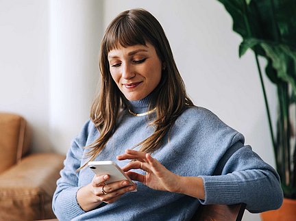Eine Frau in einem blauen Pullover, die lächelnd ihr Smartphone benutzt und drinnen in der Nähe einer Pflanze sitzt.