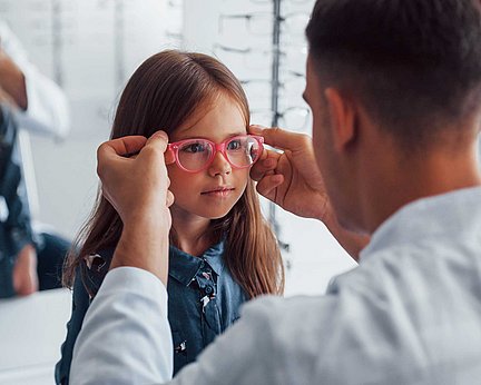 Ein Optiker passt einem jungen Mädchen in einer Augenklinik eine rosa Brille an.