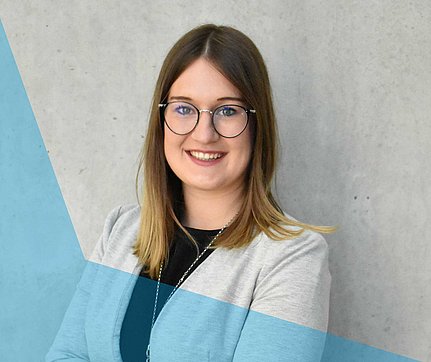 Eine lächelnde Frau mit Brille und schulterlangem Haar, die einen hellen Blazer trägt, steht vor einem grauen Hintergrund.