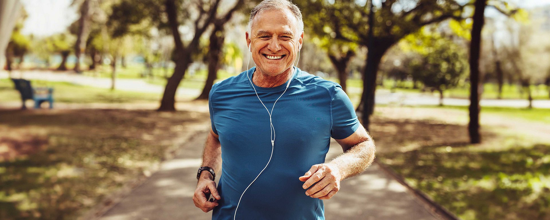 Ein lächelnder älterer Mann, der an einem sonnigen Tag in einem Park joggt; er trägt ein blaues Hemd und Kopfhörer.