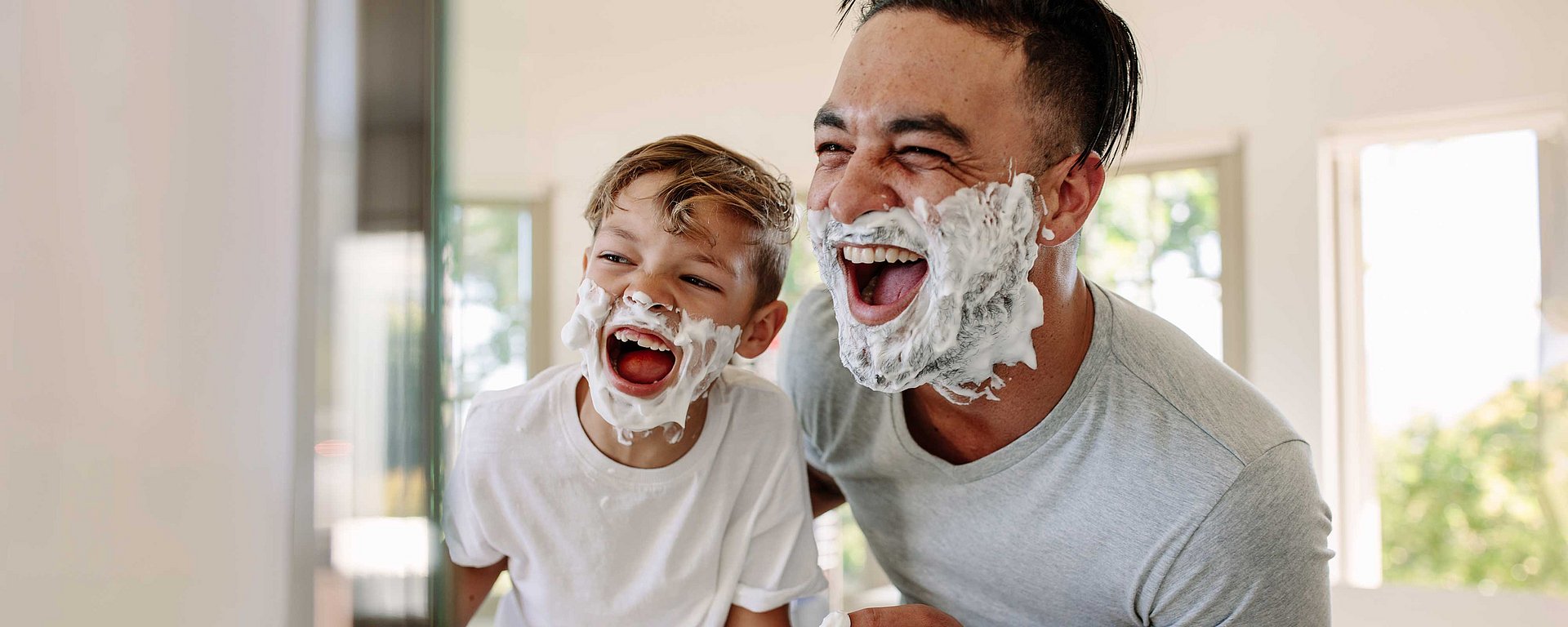 Vater und Sohn, die Rasiercreme im Gesicht haben und gemeinsam in einem hellen Badezimmer lachen.