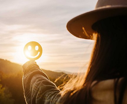 Eine Person, die eine Smiley-Ausschnittfigur in Richtung Sonnenuntergang hält, wodurch ein fröhlicher Silhouetteneffekt entsteht.