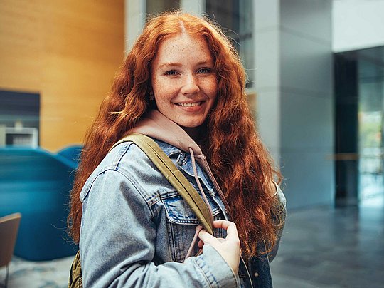Eine lächelnde junge Frau mit langen roten Haaren und einem Rucksack, die in einem modernen Gebäude steht.