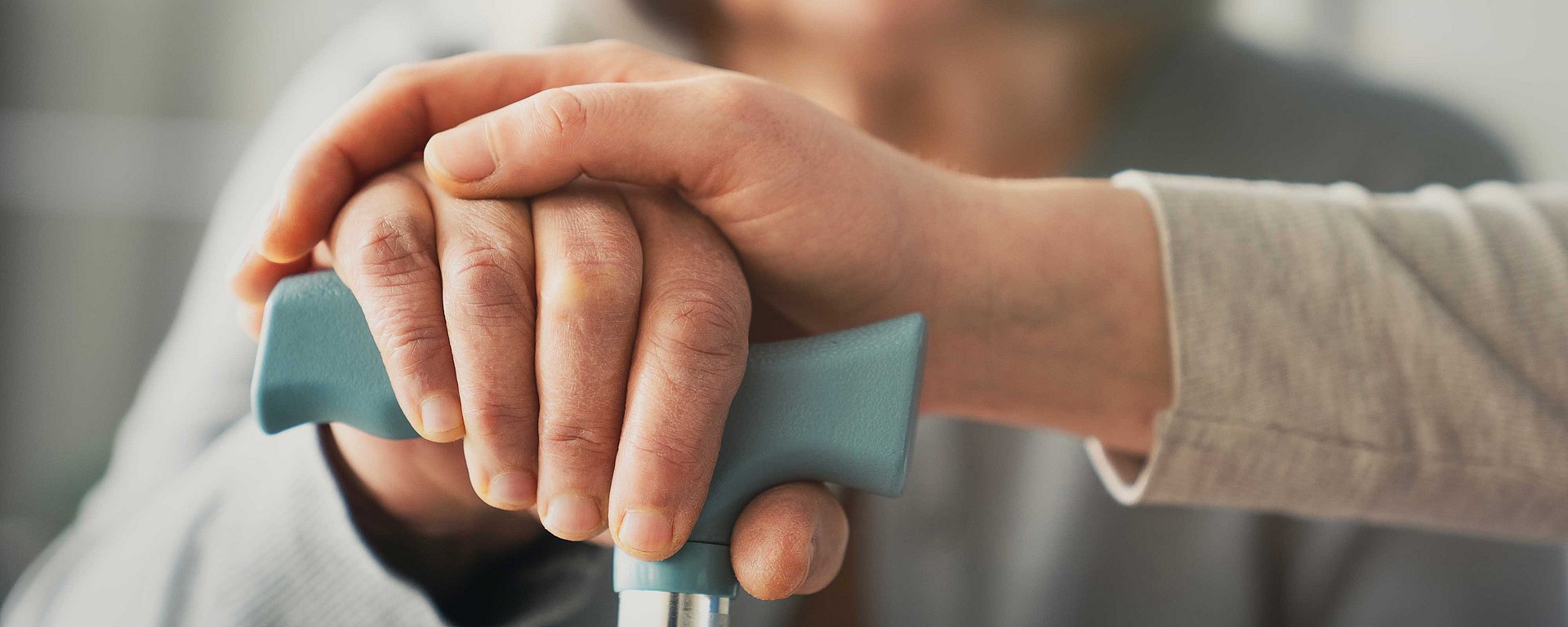 Nahaufnahme einer jüngeren Hand, die sanft auf der Hand einer älteren Person ruht, die einen Gehstock hält.