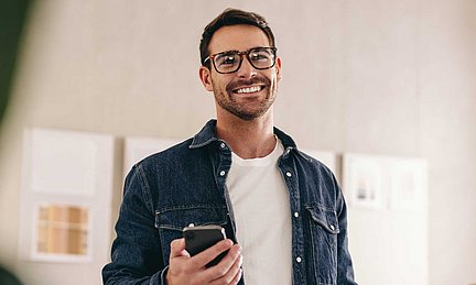 Ein lächelnder Mann mit Brille, der ein Smartphone in der Hand hält und eine Jeansjacke sowie ein weißes T-Shirt trägt, in einem Innenraum.