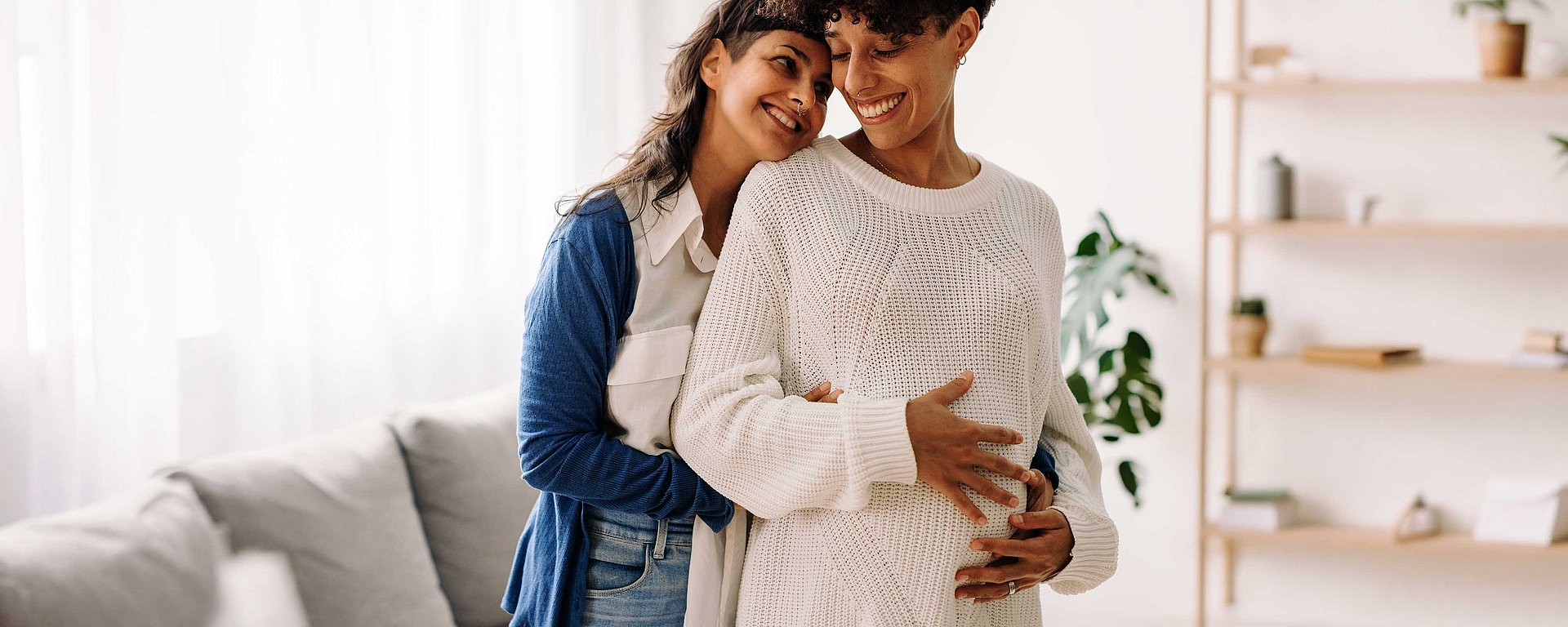 Zwei Frauen lächeln, während die eine die andere von hinten umarmt und dabei sanft ihren schwangeren Bauch berührt – in einem gemütlichen Wohnzimmer.