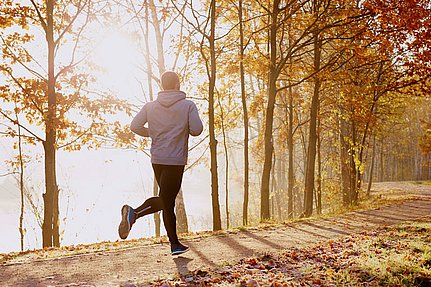 Eine Person joggt auf einem sonnendurchfluteten Waldweg, an dessen Rändern und auf dem Boden Herbstlaub liegt.