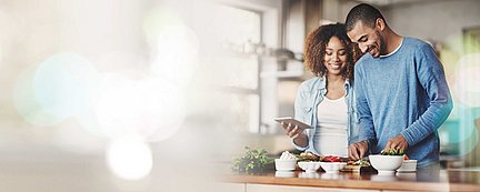 Ein lächelndes Paar, das gemeinsam in einer Küche Essen zubereitet, wobei einer ein Tablet in der Hand hält und der andere Gemüse schneidet.