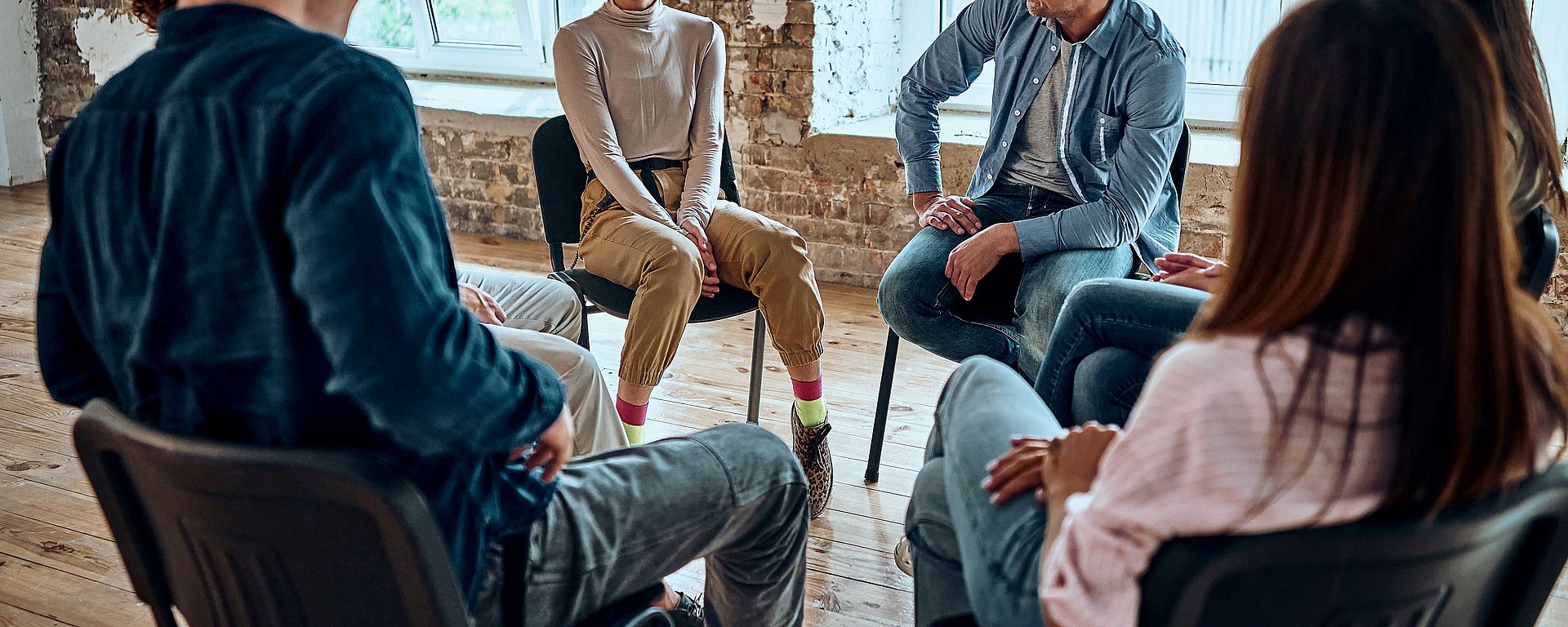 Menschen, die auf Stühlen im Kreis sitzen und in einem hellen Raum mit Holzboden eine Gruppendiskussion führen.