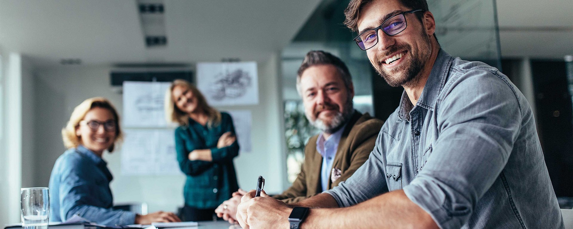 Vier lächelnde Kollegen sitzen und stehen gemeinsam in einem hellen Büro und wirken freundlich und teamorientiert.