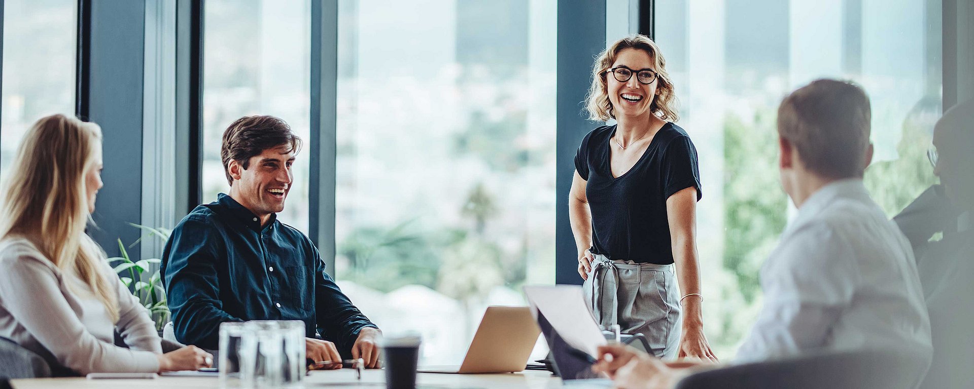 Vier Kollegen in einem modernen Büro bei einer Besprechung; eine Frau steht lächelnd da, während die anderen sitzen und zuhören.