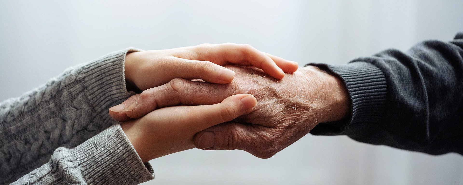 Junge Hände, die sanft die Hand einer älteren Person halten und ihr vor einem sanften Hintergrund Trost und Halt spenden.