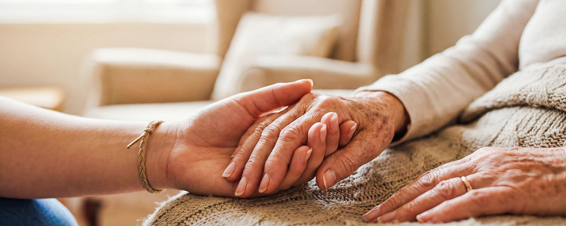 Eine jüngere Person hält sanft die Hand einer älteren Person, die auf einem mit einer Decke bedeckten Sofa sitzt.