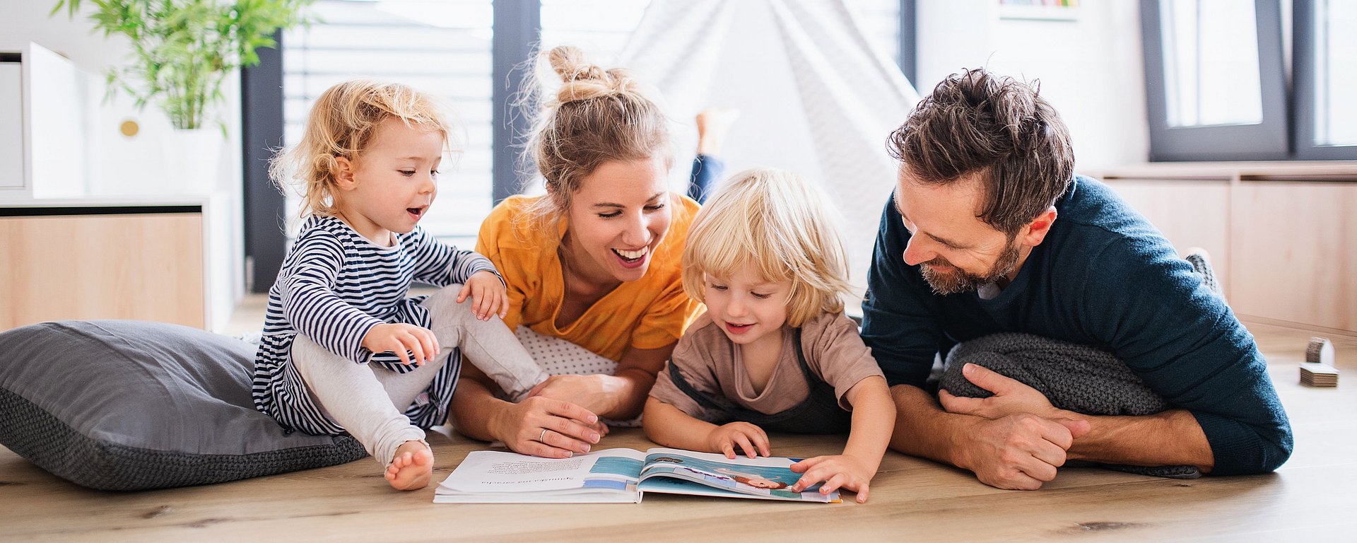 Eine vierköpfige Familie liegt auf dem Boden, lächelt und liest gemeinsam ein Buch in einem hellen Zimmer.