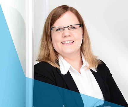 Lächelnde Frau mit Brille in Business-Kleidung, die mit verschränkten Armen vor einem weißen Hintergrund steht.