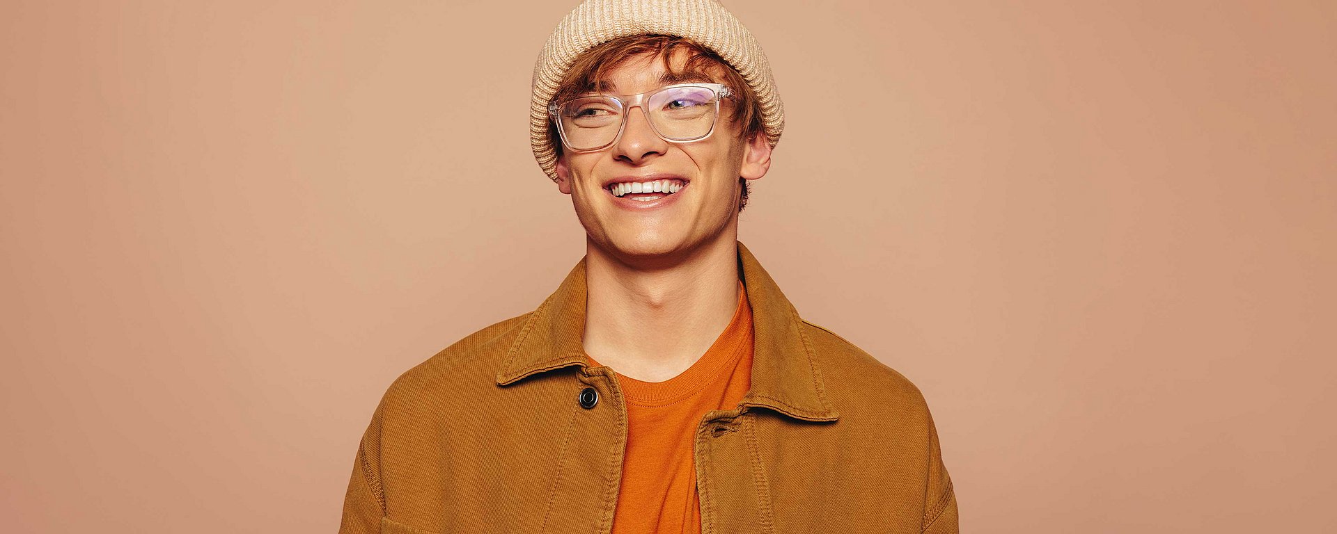 Ein junger Mensch mit Brille, Mütze und brauner Jacke lächelt vor einem schlichten beigen Hintergrund.