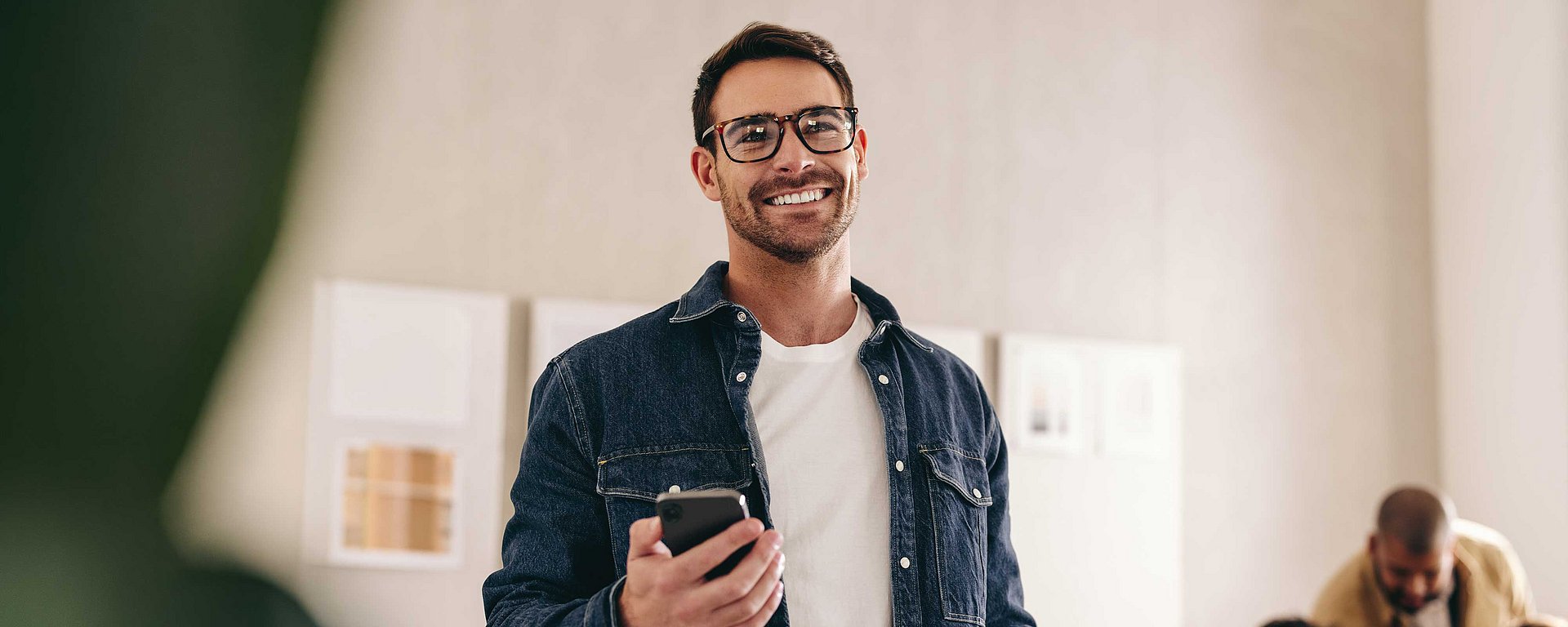 Ein lächelnder Mann mit Brille, der ein Handy in der Hand hält und in einer ungezwungenen Umgebung in einem Innenraum steht.