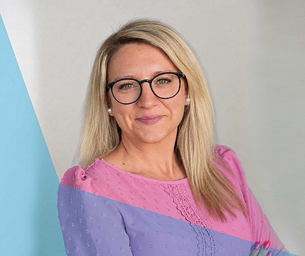 Ein professionelles Porträtfoto einer lächelnden blonden Frau, die eine Brille und ein Oberteil mit rosa-lila Muster trägt.