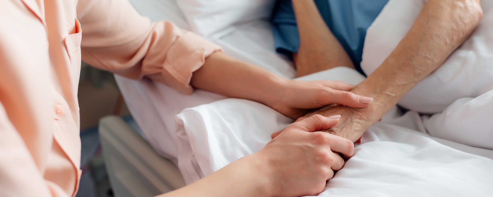 Eine Person hält sanft die Hand einer Person, die in einem Krankenhausbett liegt, und zeigt ihr damit Fürsorge und Unterstützung.