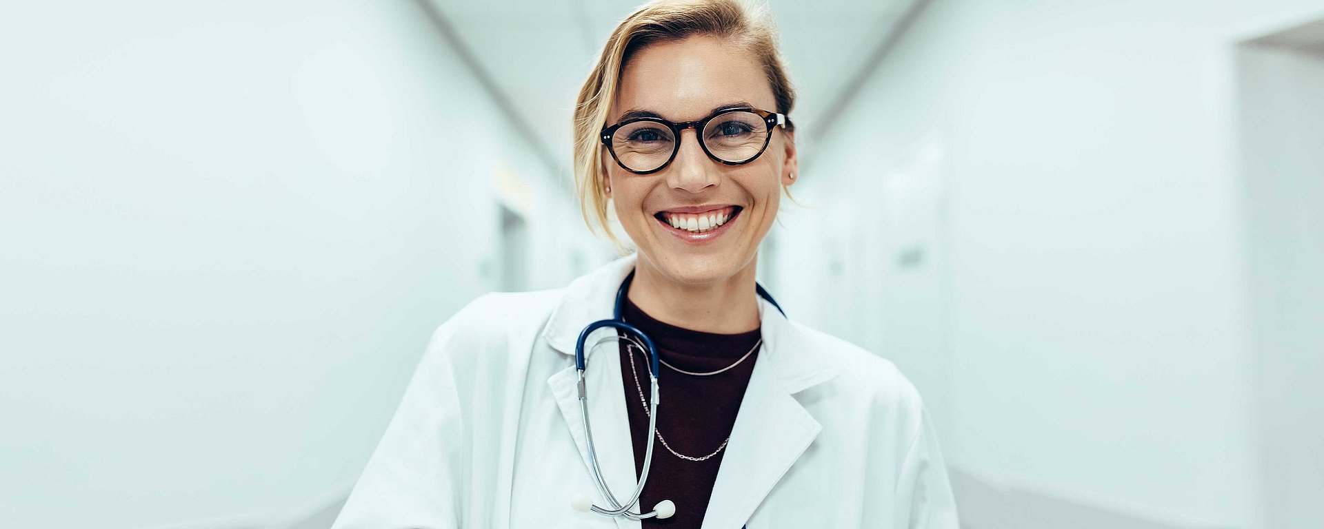 Ein lächelnder Arzt mit Brille und Stethoskop steht in einem hellen Flur und trägt einen weißen Kittel.