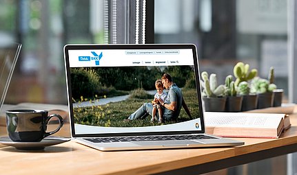Auf einem Laptop, der auf einem Schreibtisch steht, ist eine Website zu sehen, auf der ein Foto eines Mannes und eines Kindes zu sehen ist, die draußen auf dem Rasen sitzen.