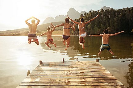 Fünf Menschen springen von einem Holzsteg in einen See, im Hintergrund sind Bäume und Berge zu sehen.
