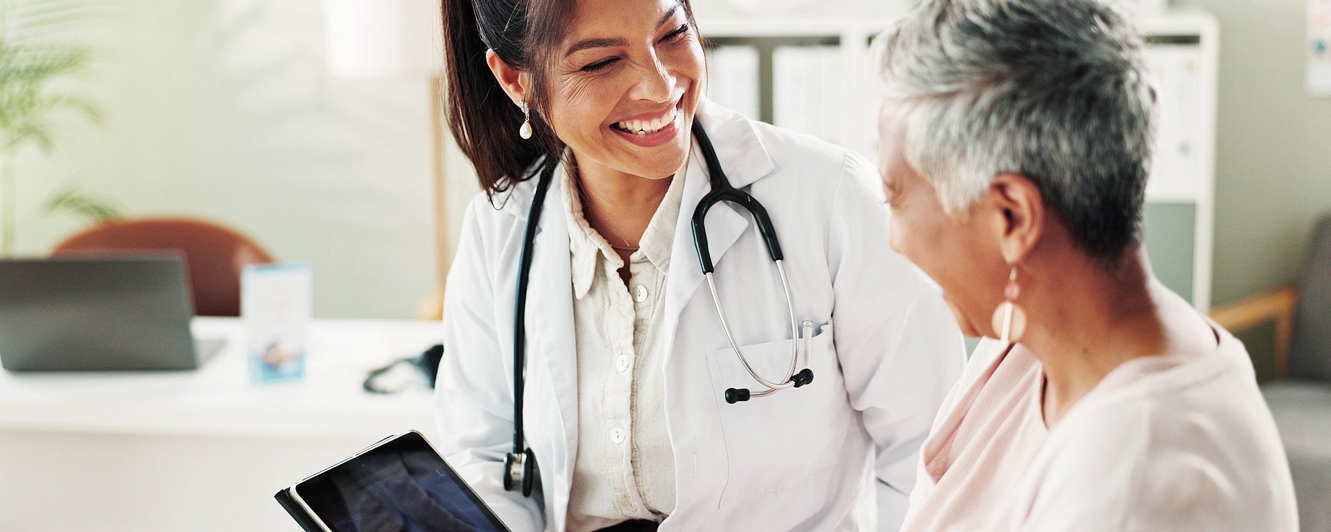 Ein lächelnder Arzt mit einem Stethoskop unterhält sich mit einer älteren Patientin; beide wirken glücklich und aufmerksam.