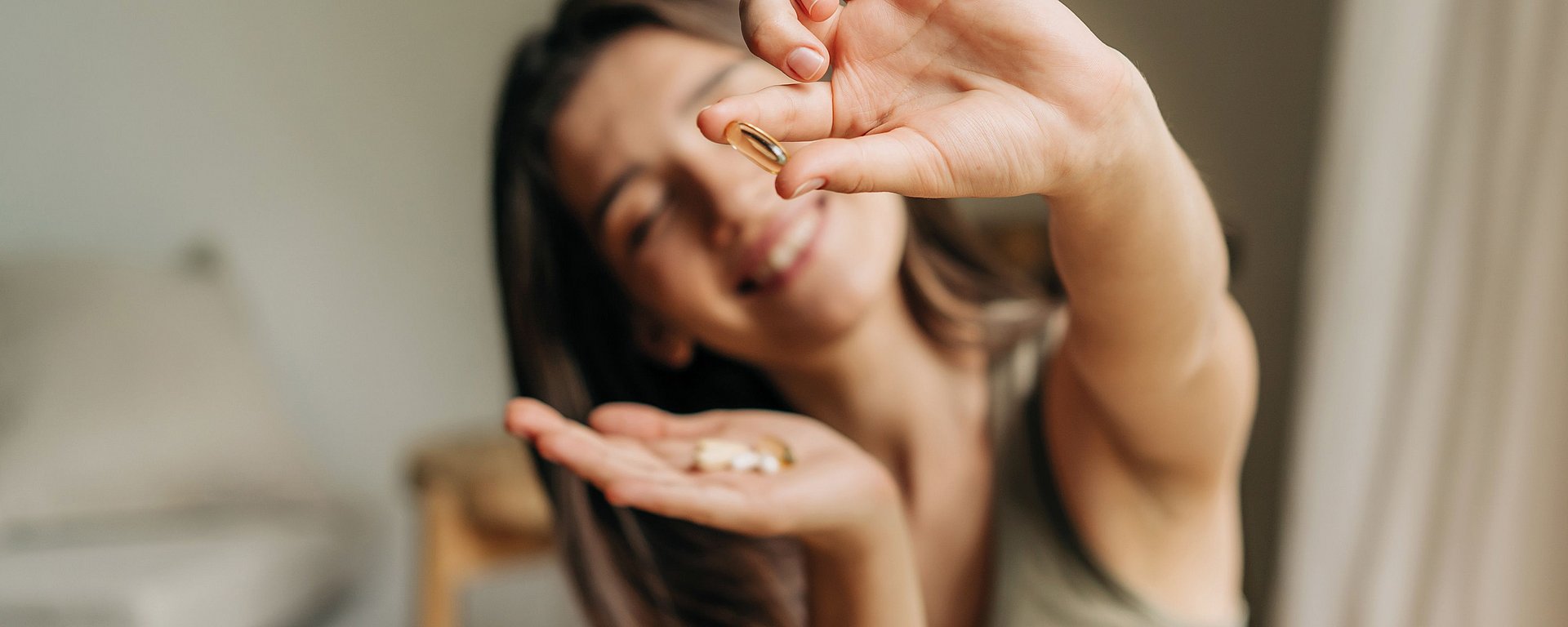 Eine lächelnde Frau, die in der einen Hand eine Kapsel und in der anderen mehrere Tabletten hält, in einem Innenraum.
