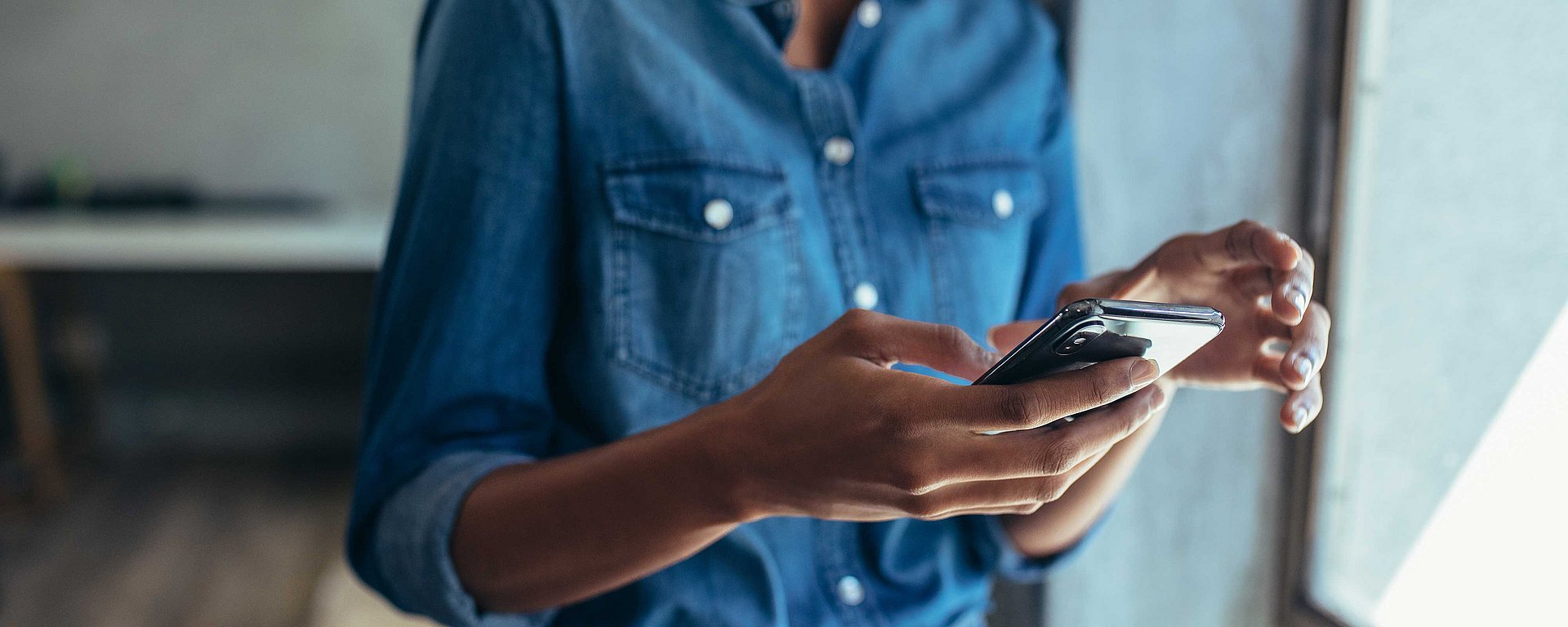 Eine Person in einem Jeanshemd, die neben einem Fenster steht und ein Smartphone benutzt.