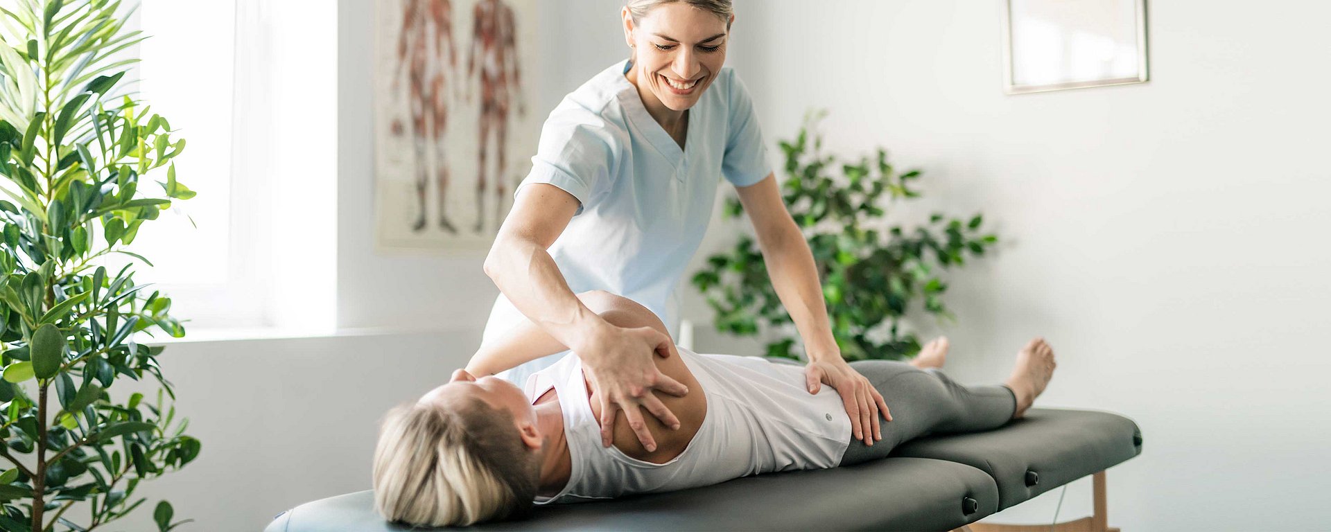 Ein Physiotherapeut hilft einer Frau auf einer Behandlungsliege in einer hellen Praxis bei einer Armdehnung.