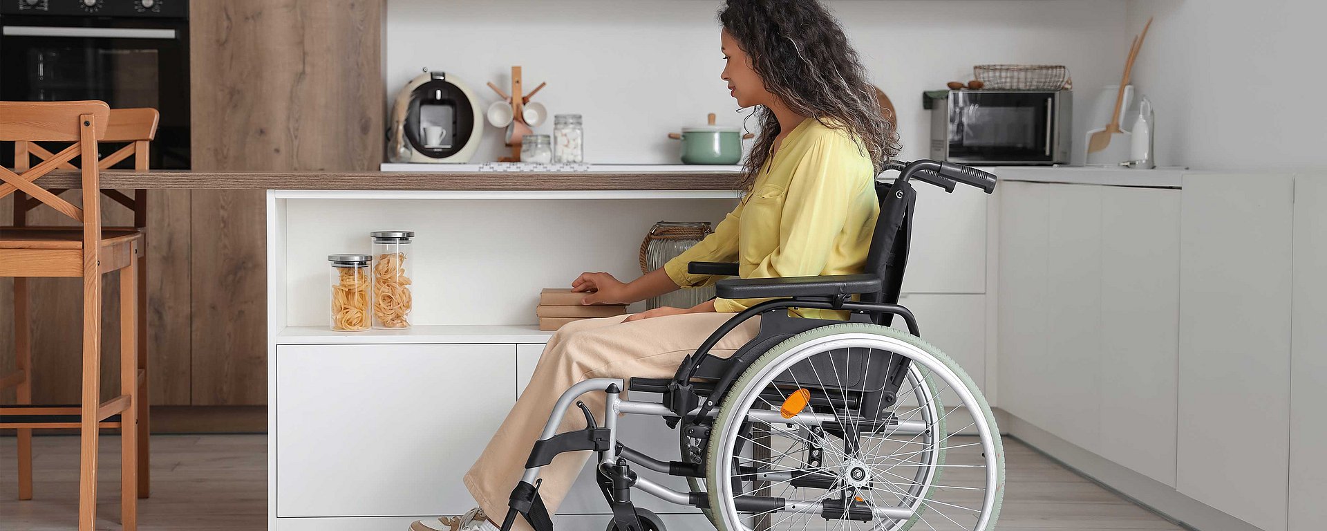 Eine Frau im Rollstuhl greift nach Gegenständen in einer modernen Küche mit weißen Schränken und Holzakzenten.