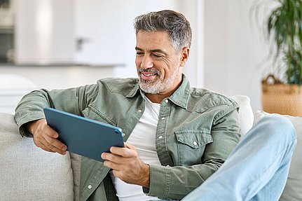 Ein lächelnder Mann sitzt auf einem Sofa und nutzt ein Tablet in einem hellen, modernen Wohnzimmer.