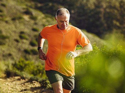 Ein älterer Mann in einem orangefarbenen T-Shirt joggt draußen auf einem Wanderweg, schaut auf seine Smartwatch und trägt Kopfhörer.
