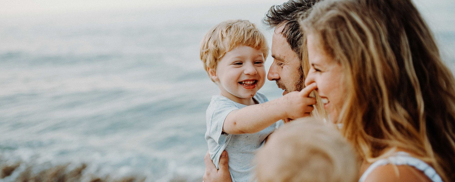 Eine lächelnde Familie mit einem kleinen Jungen und seinen Eltern, die gemeinsam die Zeit am Meer genießen.