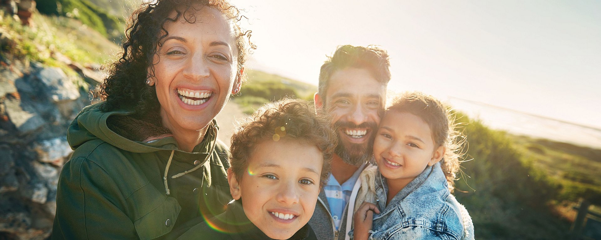 Eine lächelnde vierköpfige Familie posiert an einem sonnigen Tag im Freien, im Hintergrund sind Grünflächen und Sonnenlicht zu sehen.