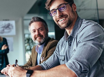 Zwei Männer sitzen an einem Tisch und lächeln; im Fokus steht ein Mann mit Brille und einem legeren Hemd.