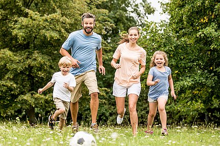 Eine lächelnde vierköpfige Familie rennt gemeinsam draußen auf dem Rasen auf einen Fußball zu.