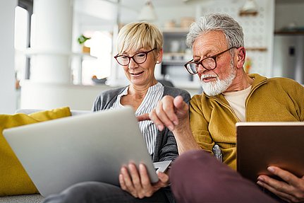 Ein älteres Paar sitzt auf einem Sofa, nutzt gemeinsam einen Laptop, wobei eine Person ein Tablet in der Hand hält.