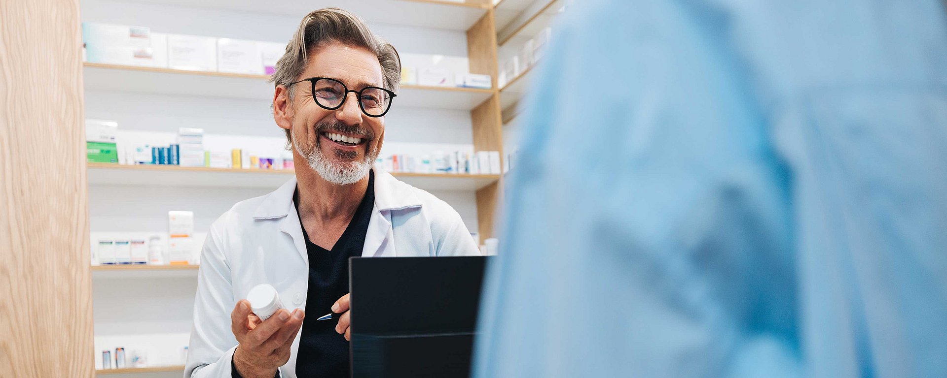 Ein lächelnder Apotheker, der eine Medikamentenflasche in der Hand hält und sich am Apotheken-Tresen mit einem Kunden unterhält.