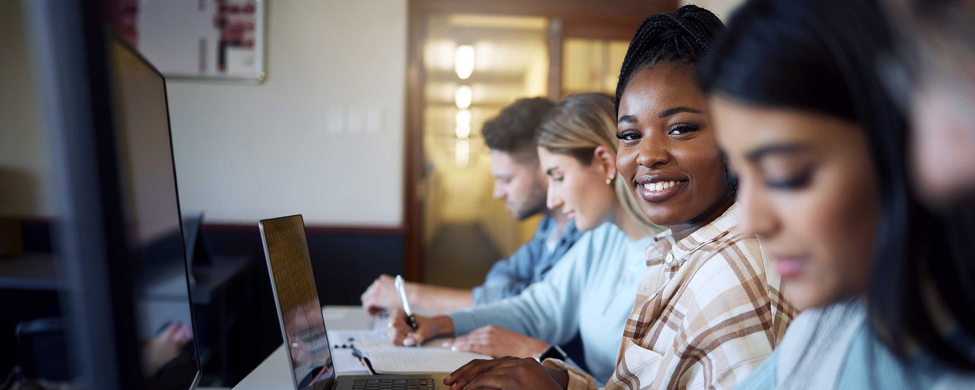 Vier Schüler sitzen in einer Reihe vor Computern, lernen und lächeln in einem Klassenzimmer.