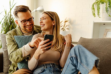 Ein lächelndes Paar sitzt auf einem Sofa und schaut sich gemeinsam ein Smartphone in einem gemütlichen Wohnzimmer an.