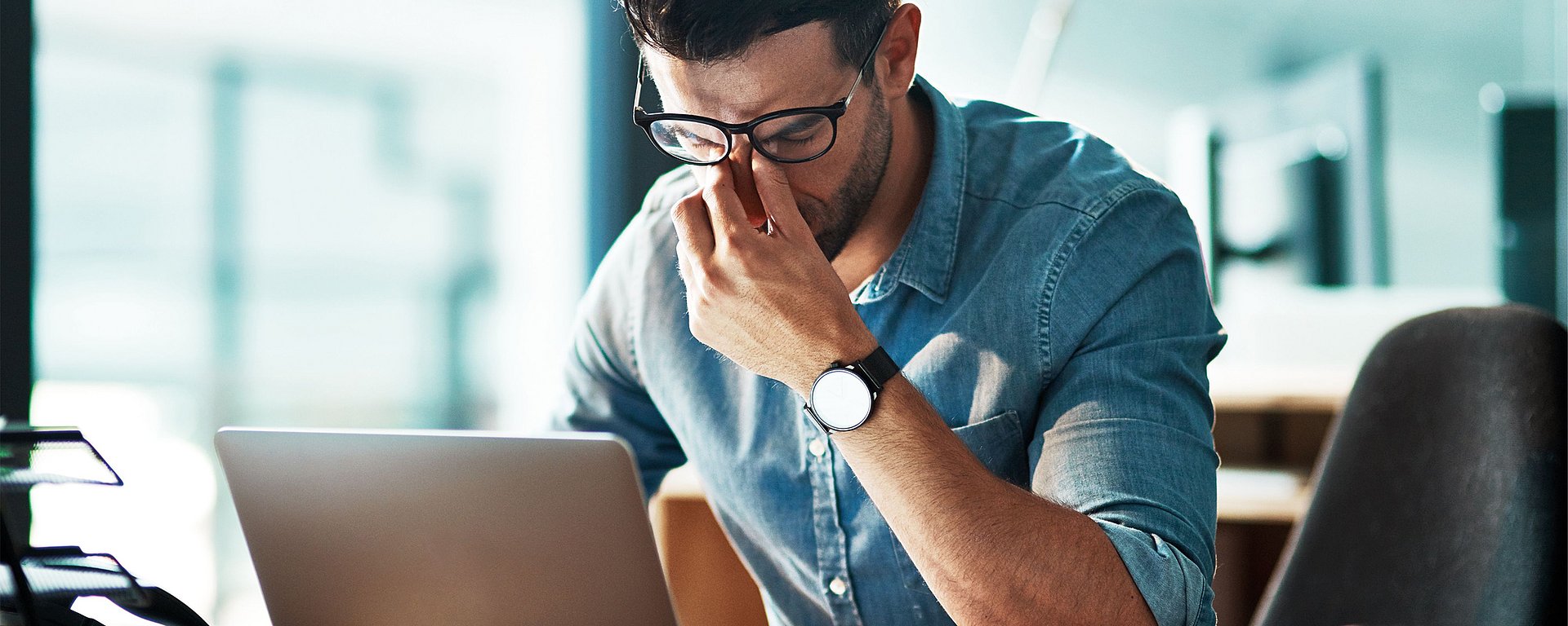 Ein Mann mit Brille sitzt vor einem Laptop, hält sich die Nasenwurzel und wirkt in einer Büroumgebung gestresst oder erschöpft.