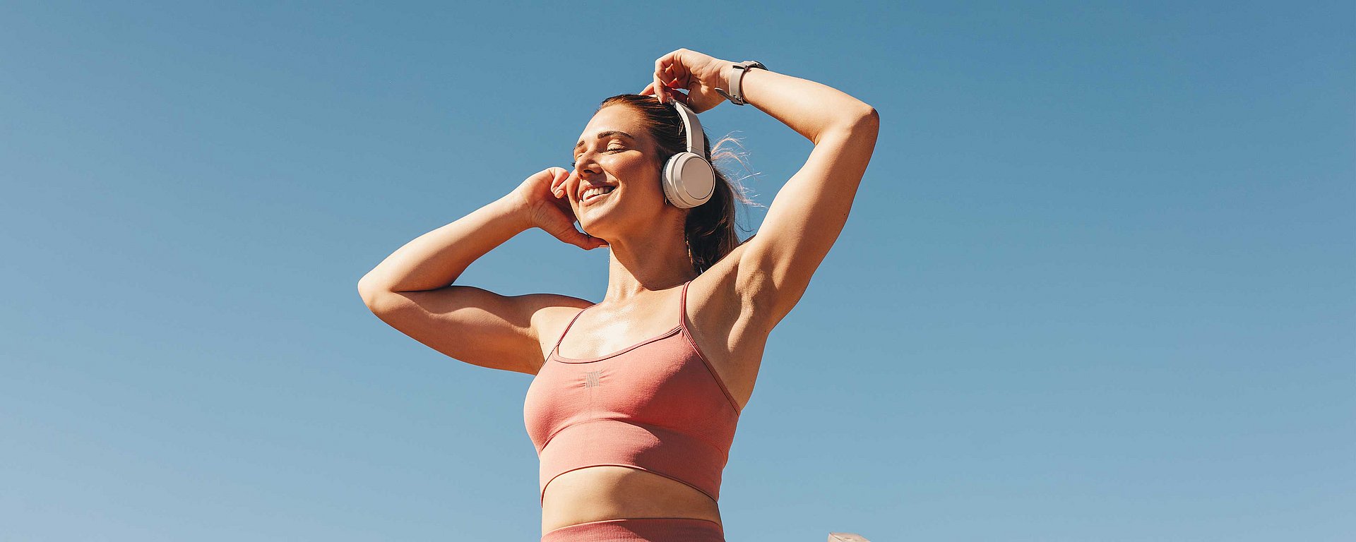 Eine Frau in rosa Sportkleidung, die mit Kopfhörern Musik hört und draußen unter einem strahlend blauen Himmel steht.