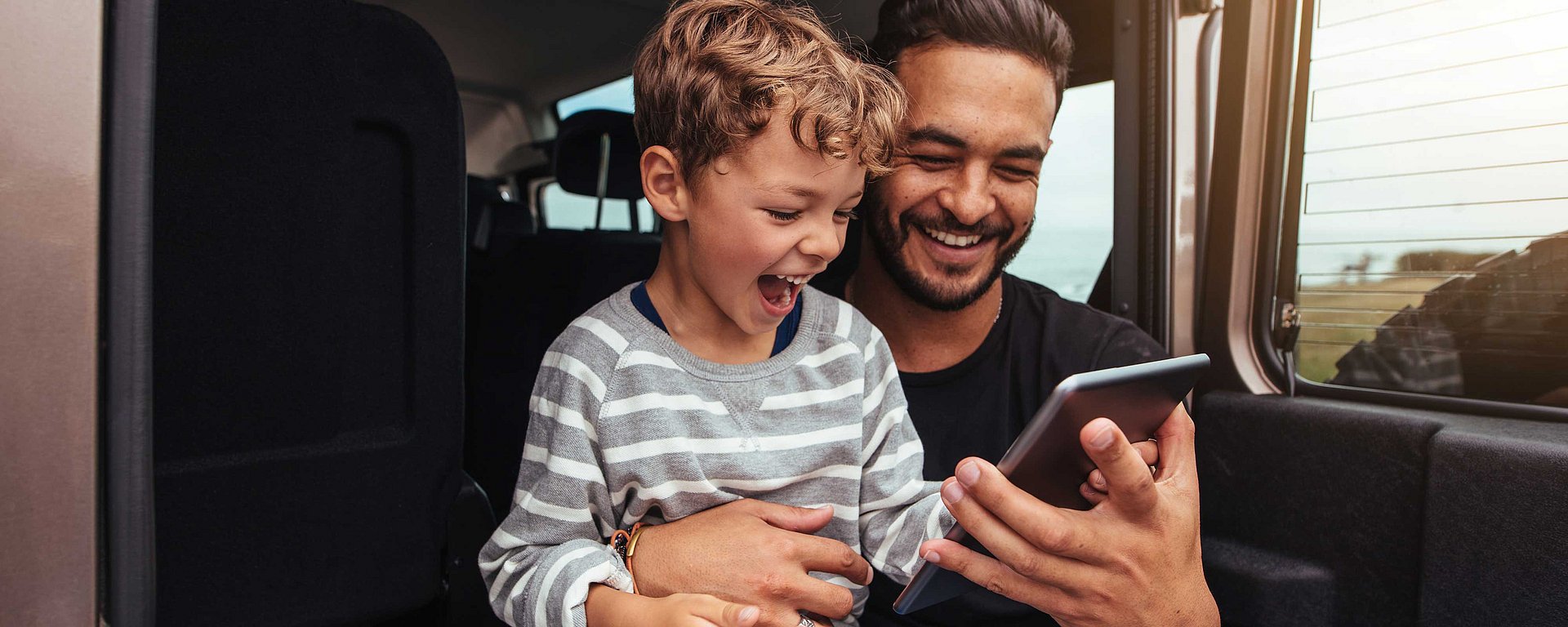 Ein lächelnder Mann und ein begeistertes Kind sitzen in einem Auto und schauen sich gemeinsam ein Tablet an.