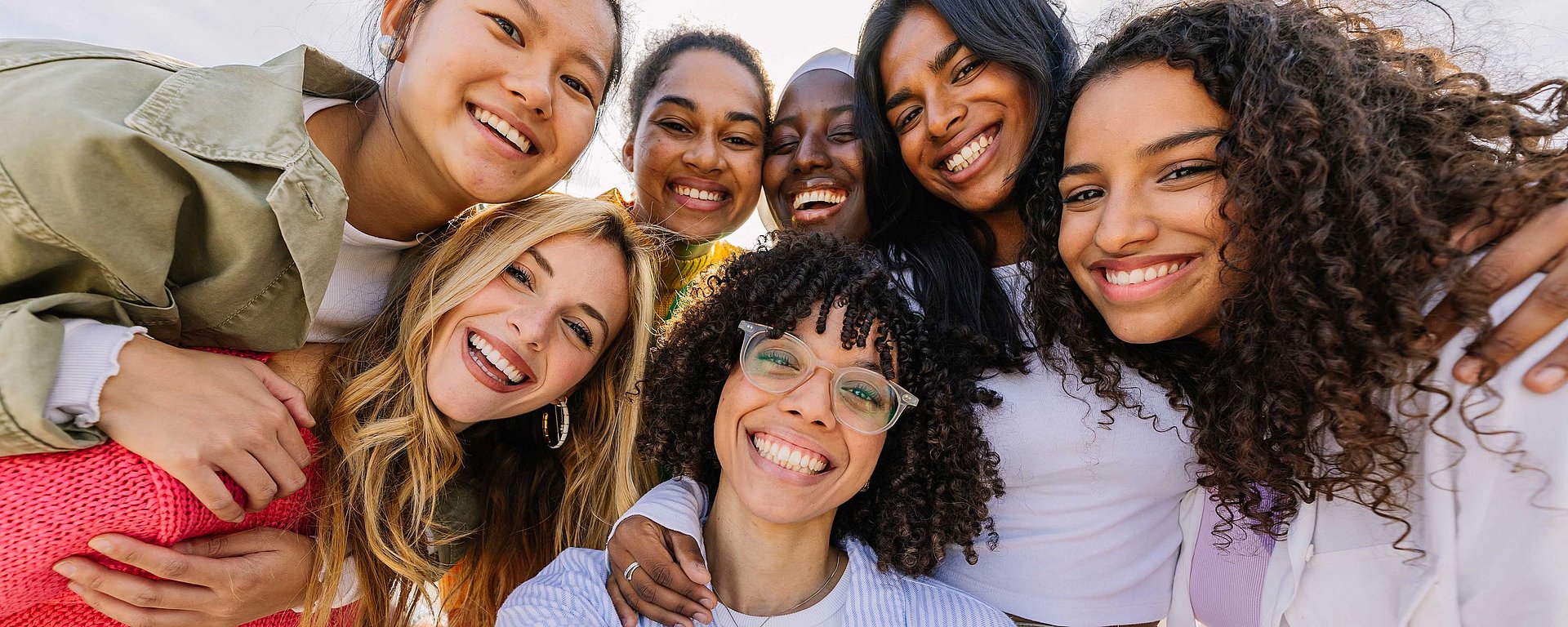 Eine Gruppe von sieben jungen Frauen, die lächelnd und eng beieinander stehend im Freien für ein Selfie posieren.