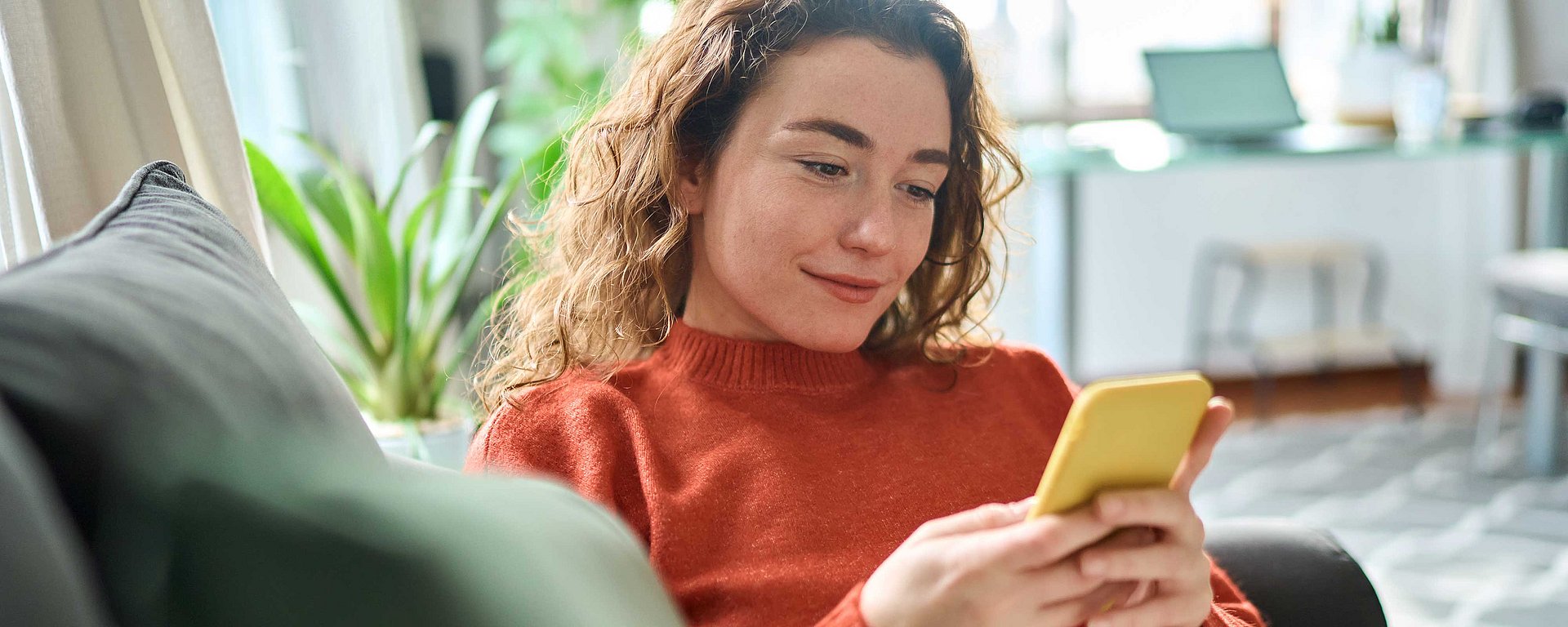 Eine Frau mit lockigem Haar in einem roten Pullover sitzt zu Hause auf dem Sofa, schaut auf ihr gelbes Handy und lächelt.