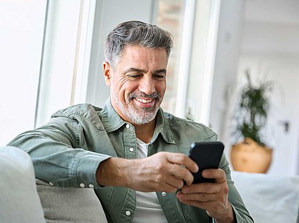 Ein lächelnder Mann mit grauen Haaren sitzt auf einem Sofa und nutzt sein Smartphone in einem hellen Wohnzimmer.